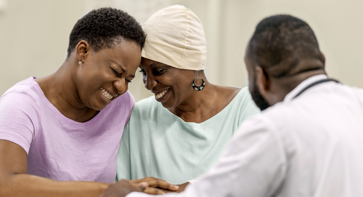 Two women, one wearing a headscarf, embrace while a doctor listens.